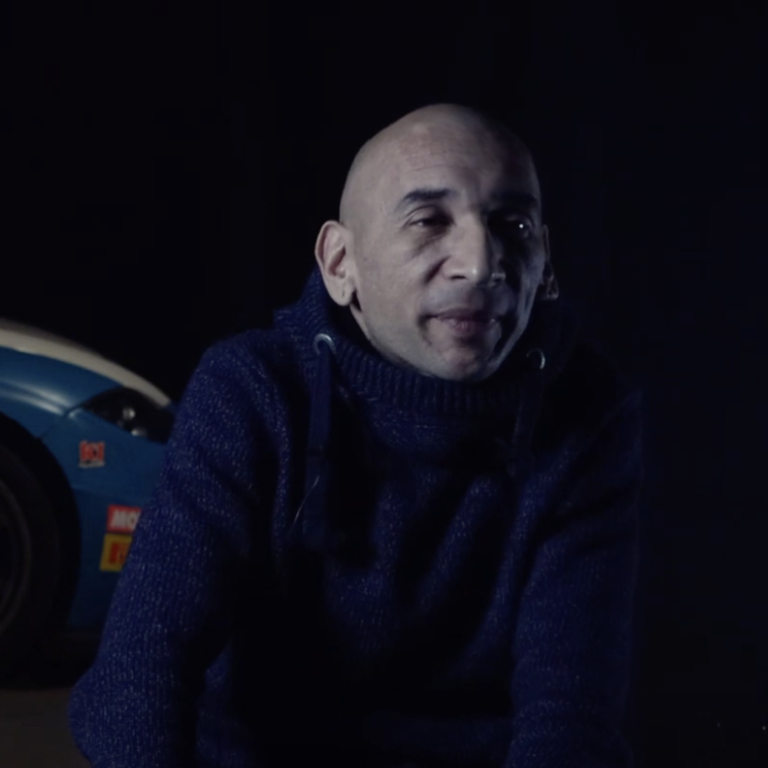 Homme chauve en pull, assis avec une voiture de course floue en arrière-plan.