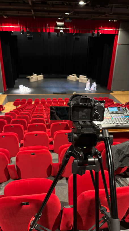 Scène de théâtre avec des sièges rouges et une caméra sur trépied au premier plan.