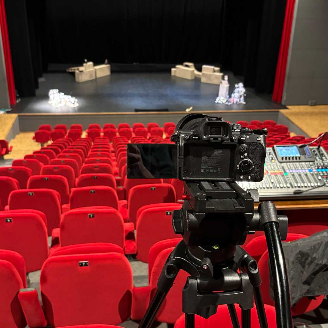Vue d'une scène de théâtre depuis des sièges rouges, avec une caméra sur un trépied.